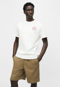 Άνδρας που φοράει λευκό t-shirt North Face και χακί σόρτς, στέκεται με τα χέρια στις τσέπες μπροστά σε απλό φόντο.