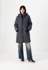 Manteau matelassé, gris foncé, avec capuche, fermeture éclair à l'avant et deux poches latérales. Présente un motif en chevrons pour la texture et le design.