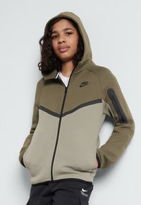 Felpa con zip Nike in verde oliva e beige. Presenta un cappuccio, accenti neri in contrasto, tasche laterali e una piccola tasca con zip sulla manica.