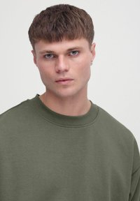 Olivzaļš džemperis ar apaļu kakla izgriezumu, mīkstu tekstūru un smalkām šūšanas detaļām. Modelim ir īsi mati un zilas acis.