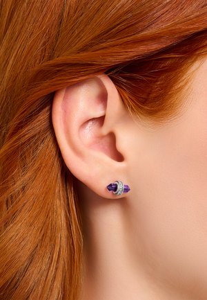 Lila Kristall Ohrringe mit silbernen Akzenten, die ein geometrisches Design und funkelnde Details aufweisen, präsentiert am Ohr eines Models.