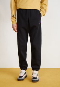 Black sweatpants with elastic cuffs, featuring a yellow logo detail. Paired with multicolored high-top sneakers and a yellow long-sleeve shirt.