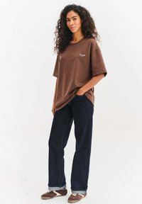 Žena s kudrnatými vlasy, oblečená v hnědém oversized tričku s malým bílým motivem řasy, tmavých džínách a hnědých teniskách, stojící s rukou v kapse.