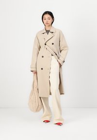 Beige, dupla gombos trench kabát fekete gombokkal, egy mintás felső fölött. Krém színű nadrággal és textúrált bézs táskával párosítva. Piros cipők.