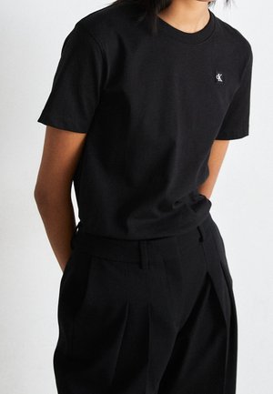 Person trägt ein eng anliegendes schwarzes T-Shirt mit kurzen Ärmeln, einem kleinen weißen CK-Logo und schwarze, hoch taillierte, plissierte Hosen.