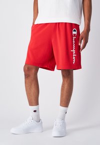Pantalones cortos deportivos rojos hechos de tela suave, con una cinturilla elástica y el logo bordado en blanco de "Champion" en el lado izquierdo.