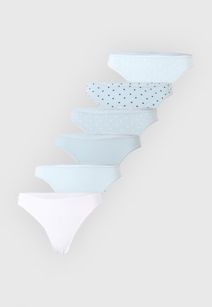 Jeune femme portant un soutien-gorge beige et une culotte bleu clair à pois, debout avec les bras derrière le dos sur un fond blanc uni.