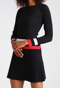 Haut noir à manches longues avec des pois blancs, doté de bandes rayées rouges et blanches au bas. Associé à une jupe noire évasée.