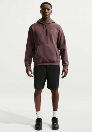 Jonge man staat, draagt een bruine Nike-hoodie, zwarte korte broek, witte Nike-sokken en zwarte sneakers tegen een effen lichte achtergrond.