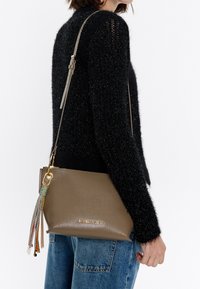 Brown textured leather handbag with gold hardware, tassel accents in multiple colors, and an adjustable strap. Styled with casual denim.