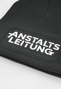Black knit beanie featuring a rectangular patch with white text "ANSTALTSLEITUNG," displaying ribbed texture and a snug fit.
