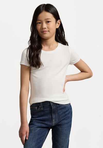 Hvit t-skjorte med korte ermer som har en liten rosa logo på brystet, kombinert med mørkeblå jeans, vist mot en ensfarget bakgrunn.