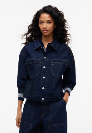 Kvinde med krøllet hår iført mørkeblå denimjakke og matchende bukser, den ene hånd i lommen, mod hvid baggrund.