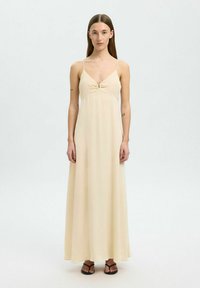 Lys beige maxi kjole med tynde stropper, en snoet frontdetalje og en glat, flydende stofstruktur. Båret med sorte flade sandaler.