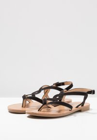 Mustat litteät sandaalit, joissa on ristikkäiset kankaiset remmit, varvasosa ja vaaleanruskea nahkapohja. Pohja on vaaleanruskea, ja siinä on hyvin vähän uritusta.