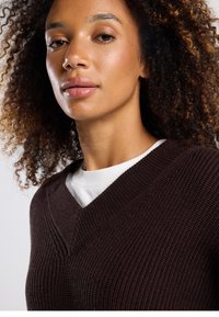 Ruskea neulottu villapaita, jossa on v-kaula-aukko ja kuvioitu kangas. Päällä valkoinen pyöreäkauluksinen paita.