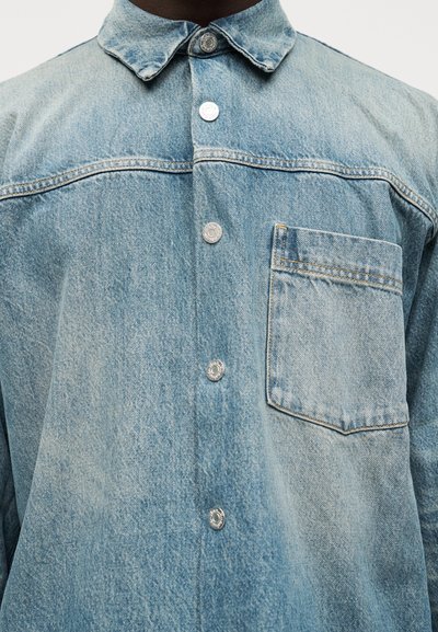 Ανοιχτό μπλε πουκάμισο από τζιν με ασημένια κουμπιά με πατητό κούμπωμα και μια μοναδική τσέπη στο στήθος, που φοριέται από ένα άτομο μπροστά σε λευκό φόντο.