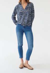 Camisetta a righe blu navy e bianche, a maniche lunghe con scollatura a V, abbinata a jeans skinny blu e scarpe beige slip-on con tacco basso.