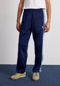 Man som bär marinblå Adidas träningsbyxor med ljusblå ränder och beige sneakers med gula ränder, stående på en blå matta.