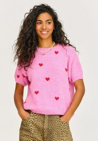 Ružový pletený sveter s krátkymi rukávmi, zdobený červenými srdiečkami. Vybavený leopardími nohavicami. Mäkká textúra a voľný strih.