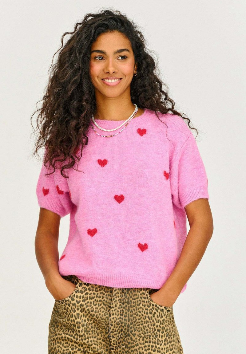 Ružový pletený sveter s krátkymi rukávmi, zdobený červenými srdiečkami. Vybavený leopardími nohavicami. Mäkká textúra a voľný strih.