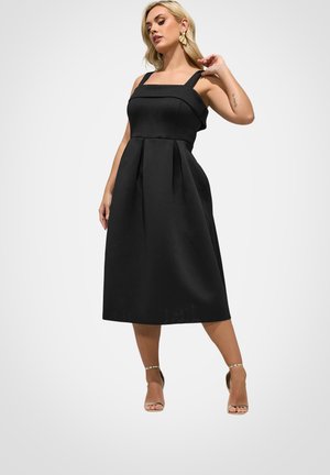 Kobieta w czarnej, bezrękawnikowej sukience midi i złotych sandałach na wysokim obcasie, pozująca z jedną ręką dotykającą włosów, a drugą przy boku.