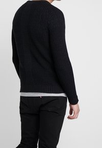 Czarny sweter robiony na drutach o prążkowanej strukturze, z okrągłym dekoltem i długimi rękawami, noszony na szarym T-shircie i czarnych spodniach.