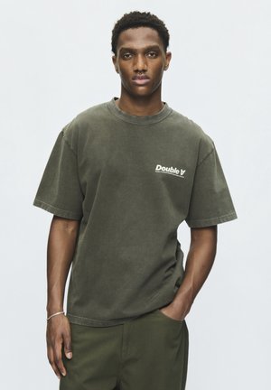 Jeune homme aux cheveux courts et bouclés portant un t-shirt Double A vert olive et un pantalon assorti, une main dans la poche, sur un fond uni.