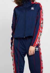 Person som bär en marinblå Adidas träningsdräkt med röda och vita blommiga ränder längs armarna och benen, med händerna avslappnat vid sidorna.