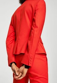 Červený na míru šitý blejzr a kalhoty, s hladkou texturou, knoflíkovými ozdobami na rukávech a strukturovaným střihem vzadu.