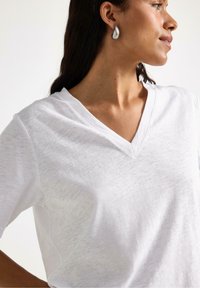 T-shirt bianco a V in tessuto leggero, con vestibilità morbida e maniche corte. Texture liscia con un leggero lucido.
