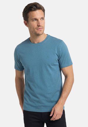 Mężczyzna z krótkimi brązowymi włosami, ubrany w niebieski t-shirt z krótkim rękawem i czarne spodnie, patrzący nieco w lewo na tle białego tła.