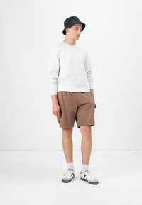 Felpa grigia, pantaloni corti marroni, cappello a secchiello nero, calzini bianchi e sneaker con accentuazioni nere. Outfit casual su uno sfondo semplice.