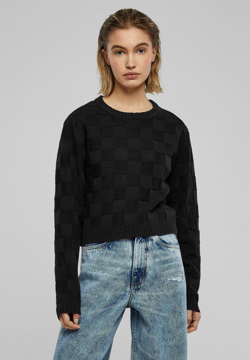 Zwarte gebreide trui met een geruit patroon, ronde halslijn en ribbelzoom, gecombineerd met lichtblauwe versleten jeans.