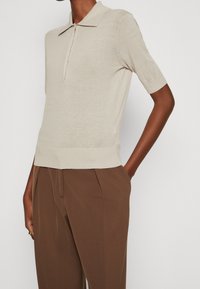 Beige Kurzarm-Strick-Polohemd kombiniert mit hochgeschnittenen braunen Faltenhosen an einer stehenden Person.