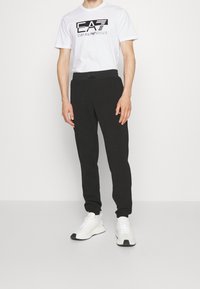 Vit kortärmad t-shirt med svart EA7-logotyp, i kombination med svarta joggers med elastisk midja. Bärs med vita sneakers.