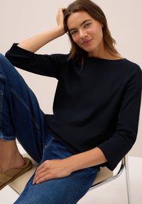 Jeune femme portant un haut noir à manches longues et un jean bleu, assise sur une chaise avec une jambe relevée et la main reposant sur sa tête.