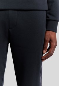 Donkerblauwe joggingbroek van een gladde stof. Heeft een hand die op de zijnaad rust, die is afgezet met een lichter streepje.