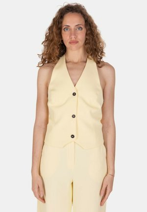 Donna con capelli ricci che indossa un gilet giallo pallido senza maniche con bottoni e pantaloni abbinati, in piedi davanti a uno sfondo chiaro e uniforme.