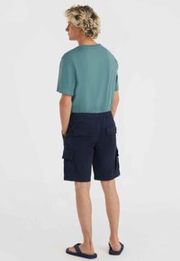 Camiseta verde de manga corta y pantalones cortos de carga en color marino con bolsillos laterales, cinturilla elástica y frente plano. Las sandalias completan el conjunto.