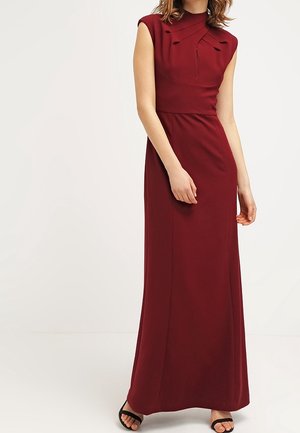Vrouw draagt een mouwloze, vloerlange bordeauxrode jurk met gevouwen details op de borst en zwarte open-toe hakken, staand tegen een effen achtergrond.