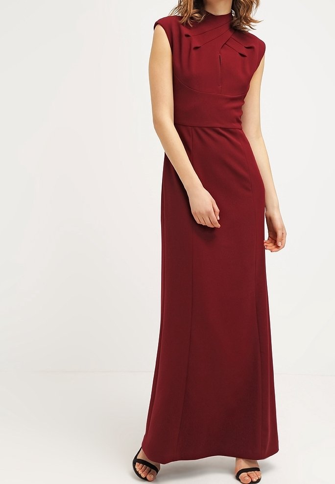 Femme portant une robe bordeaux sans manches et longue jusqu'au sol, avec des détails plissés sur la poitrine, et des talons ouverts noirs, debout devant un fond uni.