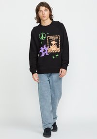 Zwarte sweatshirt met grafische prints in paars, groen en oranje, met een vredessymbool en cartoonachtige ontwerpen, gecombineerd met lichtblauwe jeans.