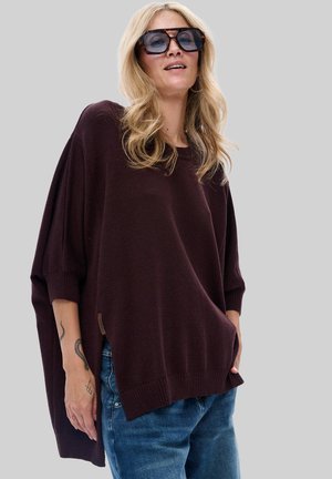 Blonde Frau, die große Sonnenbrillen trägt, einen dunklen oversized Pullover und blaue Jeans anhat, mit sichtbaren Tattoos auf ihrem linken Unterarm, steht vor einem grauen Hintergrund.
