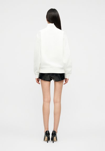 Femme aux longs cheveux foncés portant un pull à col roulé blanc, un short noir brillant et des chaussures à talons hauts noires, debout de dos.