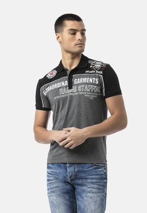 Mann in schwarzem und grauem Polohemd mit weißen Schriftzügen und Aufnähern steht mit verschränkten Händen, blickt nach links und trägt blaue Jeans.