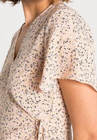 Acercamiento de una mujer que lleva un vestido envolvente floral de color rosa claro con mangas cortas y onduladas, y un detalle de lazo lateral.