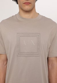 Lys beige bomulds t-shirt med rund hals, der har en tekstureret rektangulær grafik med teksten "A|X" og "ARMANI EXCHANGE BLUE EDITION".