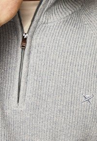 Primo piano di un maglione zip-up grigio chiaro con texture, con un piccolo logo ricamato di remi incrociati sul petto.