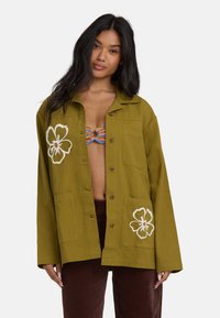 Chemise verte à manches longues avec deux poches frontales, broderies florales et coupe décontractée, fabriquée dans un tissu texturé.